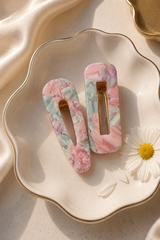Two rectangular hair claw clips with a pink, green, and white marble pattern, displayed on a scalloped cream ceramic dish with a gold rim, decorated with a white daisy flower and draped with satin fabric.