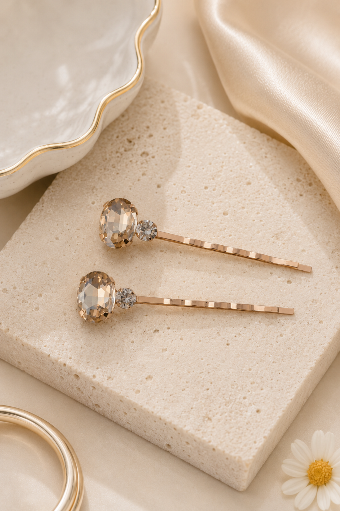 Two rose gold hair pins with clear faceted gemstones and small clear accent stones, displayed on a textured beige stone block with elegant gold-toned props.