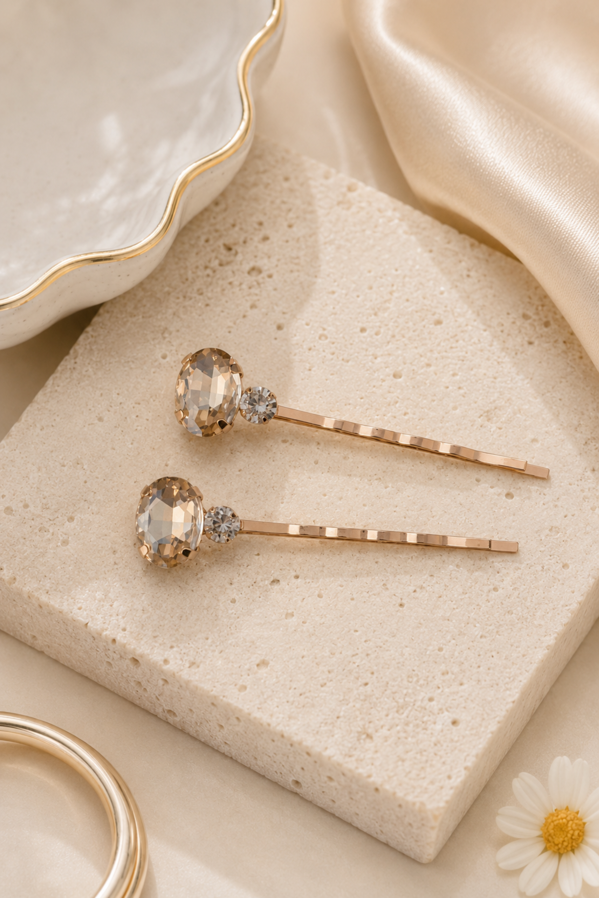 Two rose gold hair pins with clear faceted gemstones and small clear accent stones, displayed on a textured beige stone block with elegant gold-toned props.