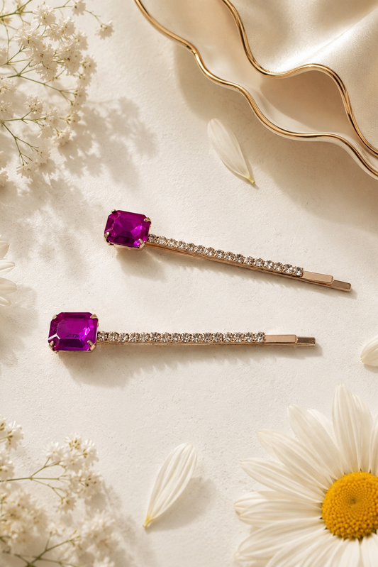 Two rose gold hair pins with rectangular purple gemstones and clear crystal-studded bars, arranged on a cream textured surface with white flowers and a gold dish.