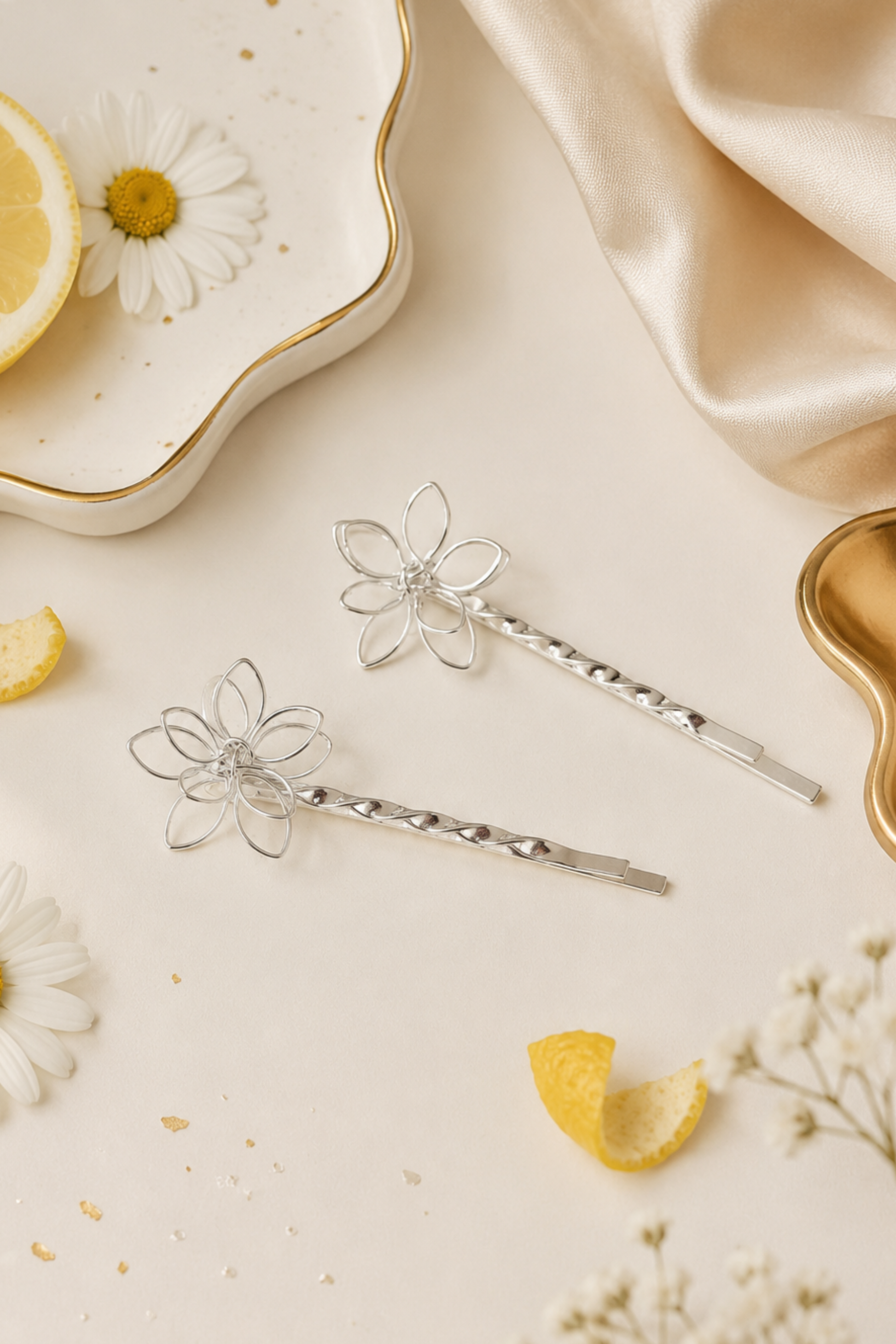 Two silver flower-shaped hair pins with twisted wire stems and small clear rhinestone centers, arranged on a cream surface with decorative daisies, lemon slices, and a gold dish.