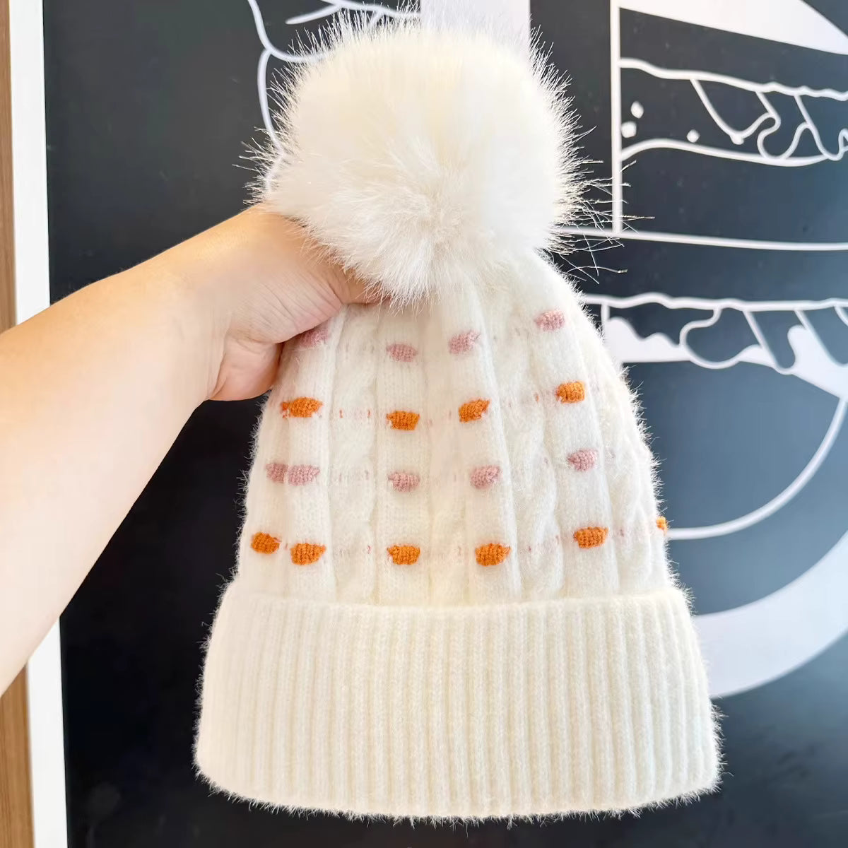 White knit beanie with a fluffy pom-pom held by a hand against a blackboard background.