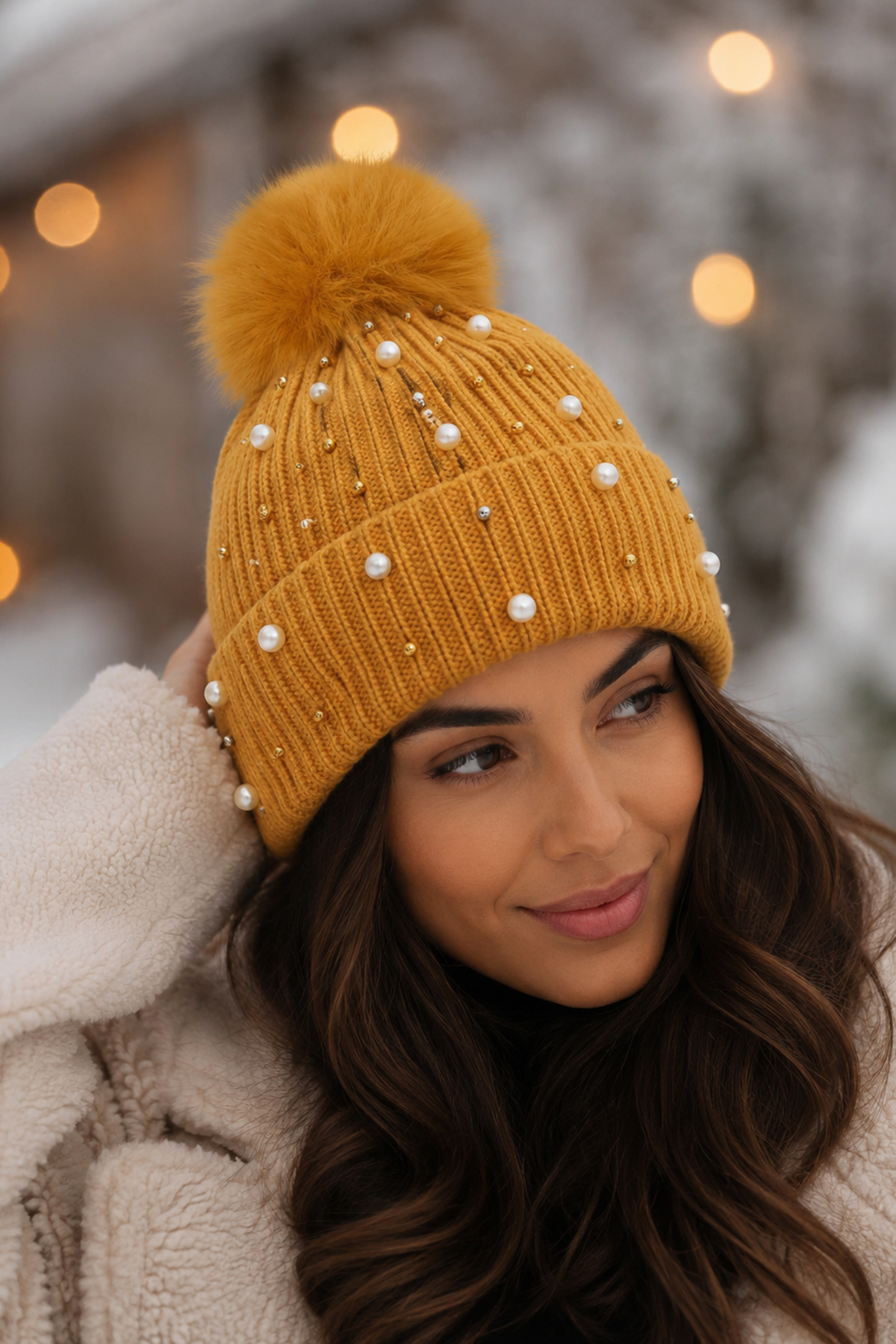 Woman wearing a mustard yellow knit beanie with a pom-pom and pearl embellishments in a snowy setting.