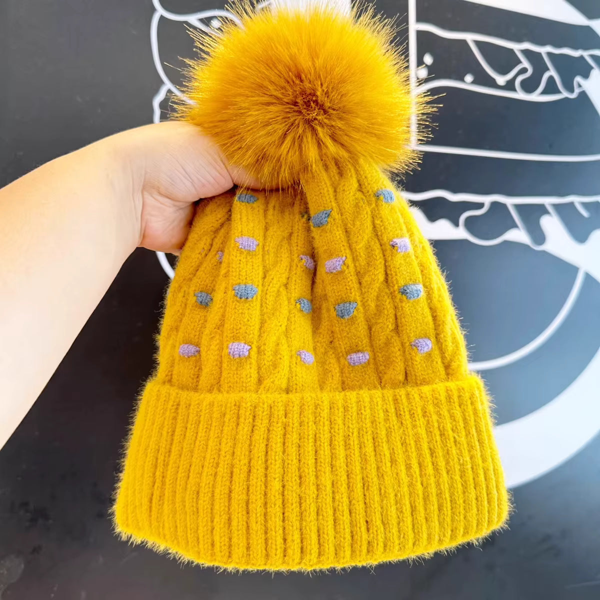Yellow knit beanie with a fluffy pom-pom held by a hand against a dark background.