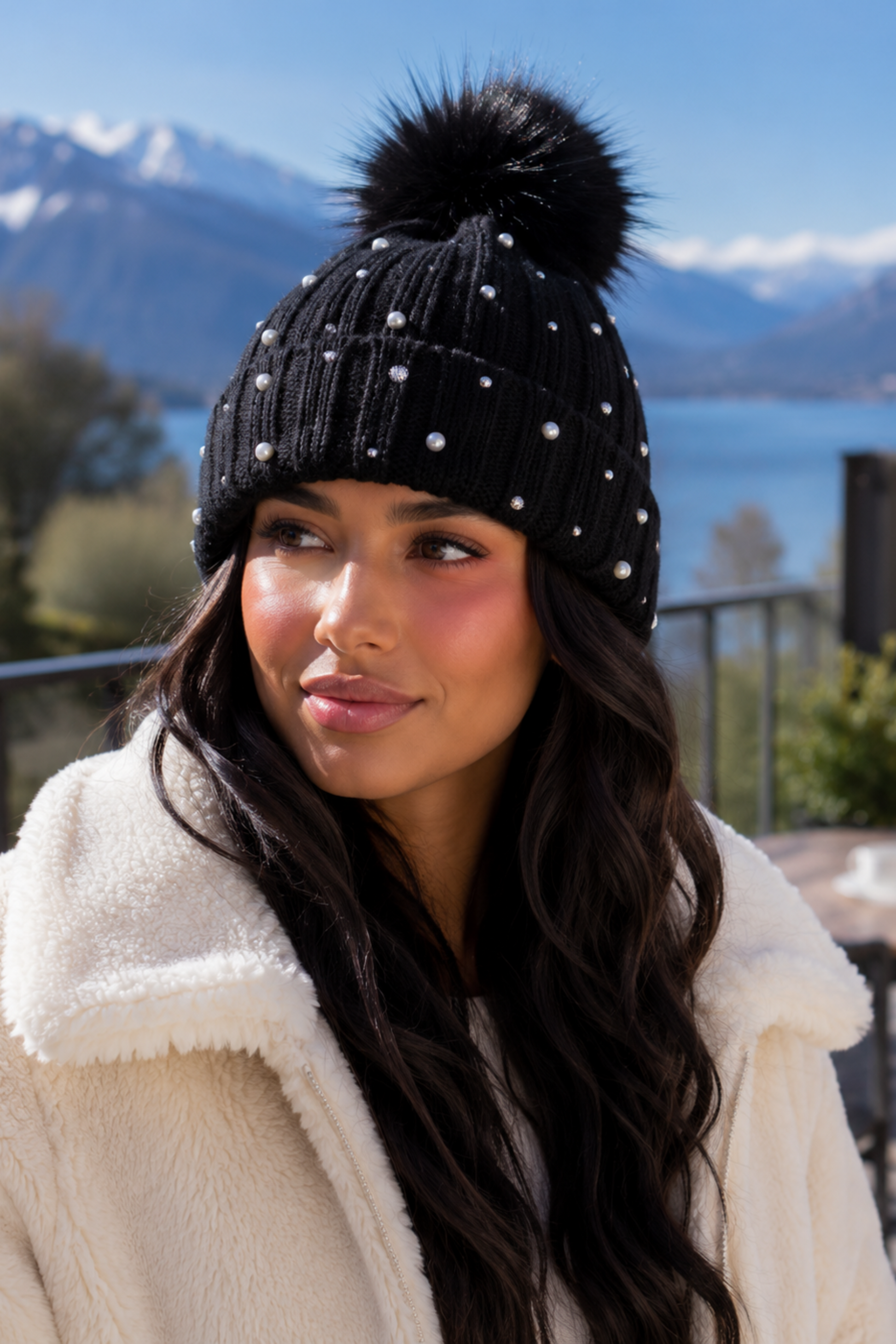 A woman wearing a black ribbed knit pom-pom beanie with white rhinestone accents and a fluffy cream sherpa jacket, photographed outdoors with mountains and a lake in the background.