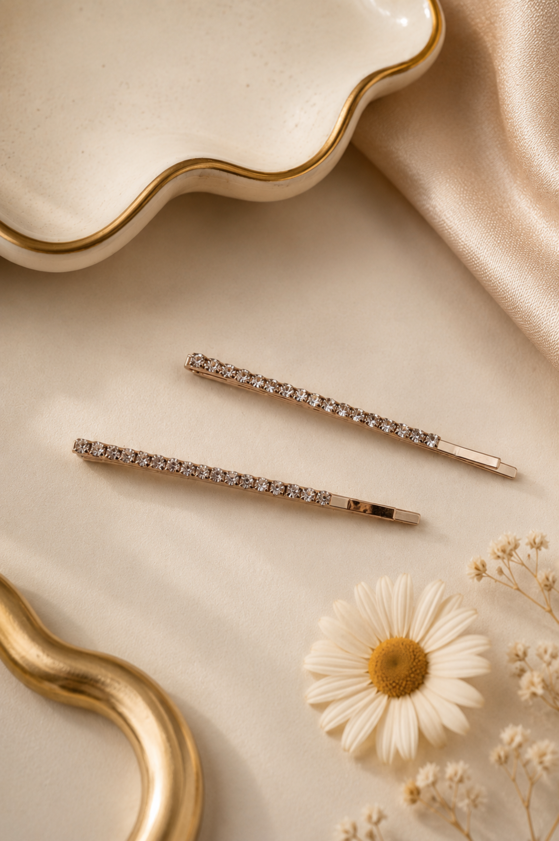 Two slim gold-toned hair pins embellished with clear crystals, arranged on a cream-colored surface with a gold dish, satin fabric, and a small daisy flower.