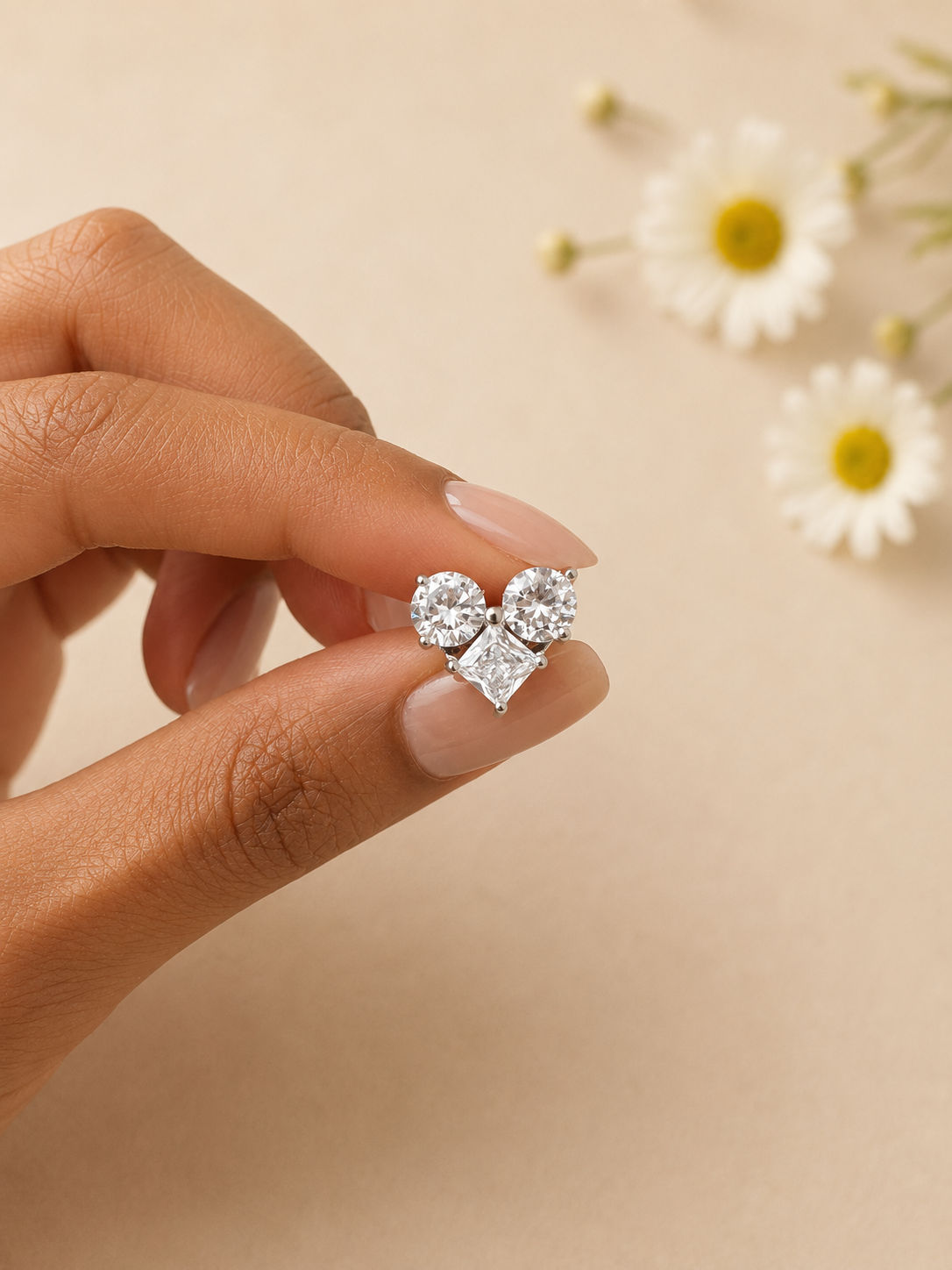 Silver earrings held between fingers with a soft beige background and flowers.
