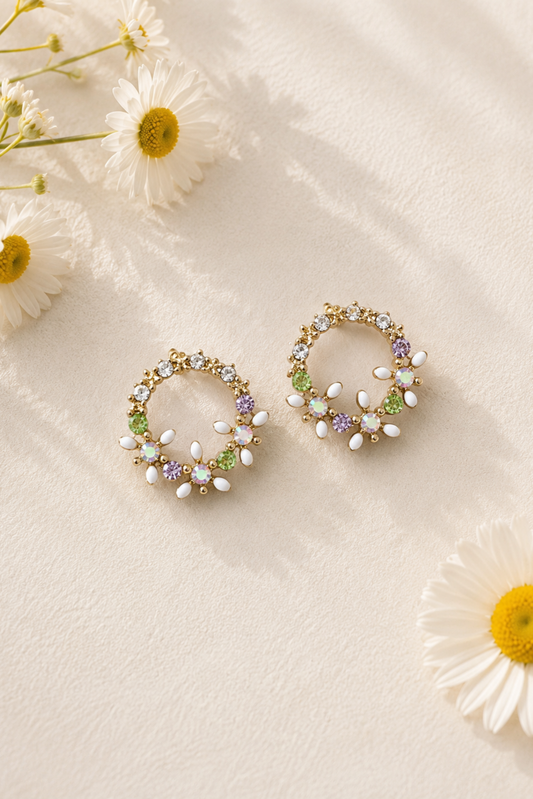 A pair of gold-tone hoop earrings decorated with white faux pearls, green and purple crystals, and clear rhinestones, styled on a textured cream background with white daisies.