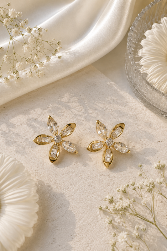 A pair of gold flower-shaped stud earrings with clear marquise-cut stones, styled on a textured cream surface with white satin, dried flowers, and a glass bowl.