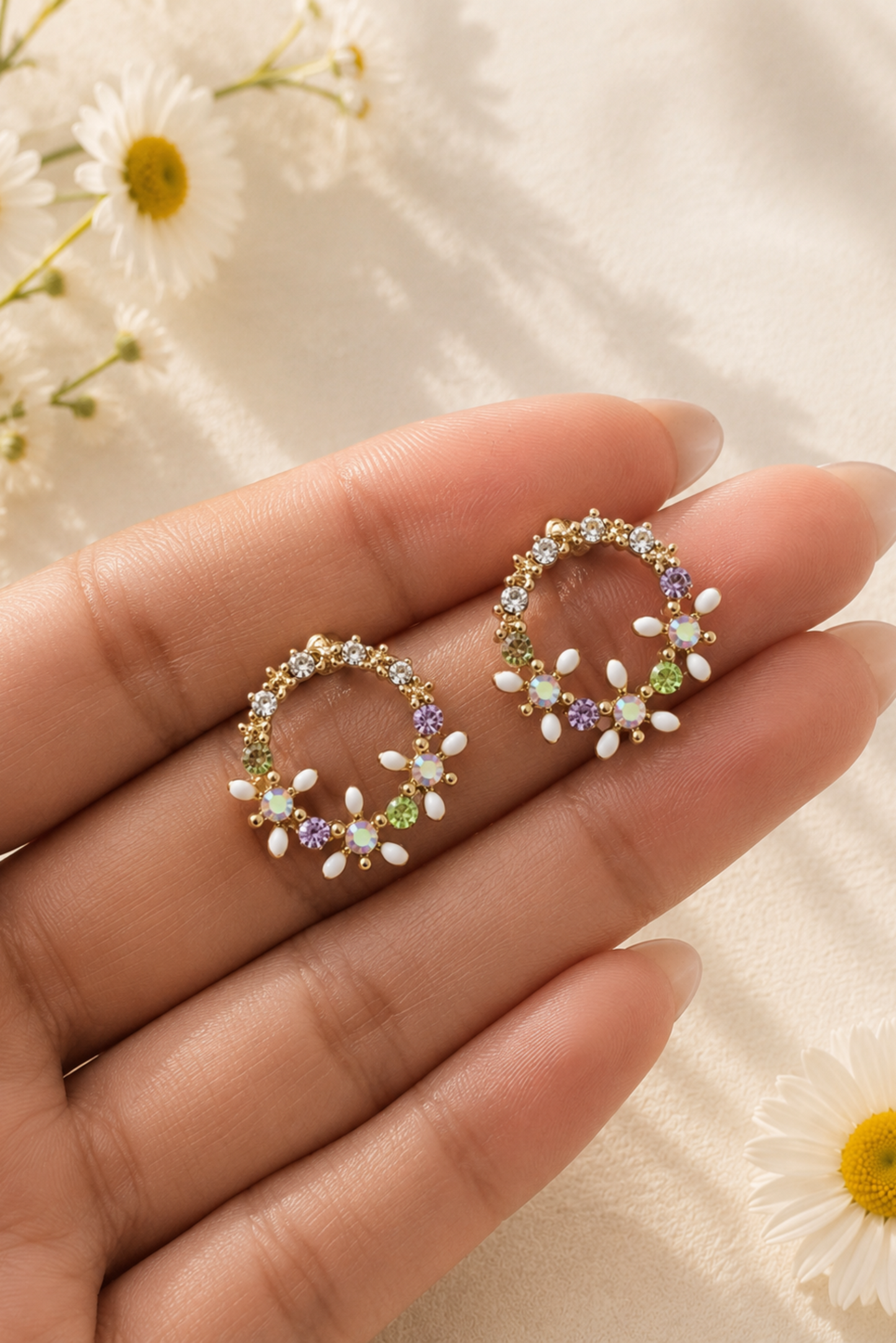 A pair of gold-toned circular stud earrings decorated with small white pearl-like beads and colorful gemstones, displayed on a person's hand against a soft cream fabric background with daisy flowers.