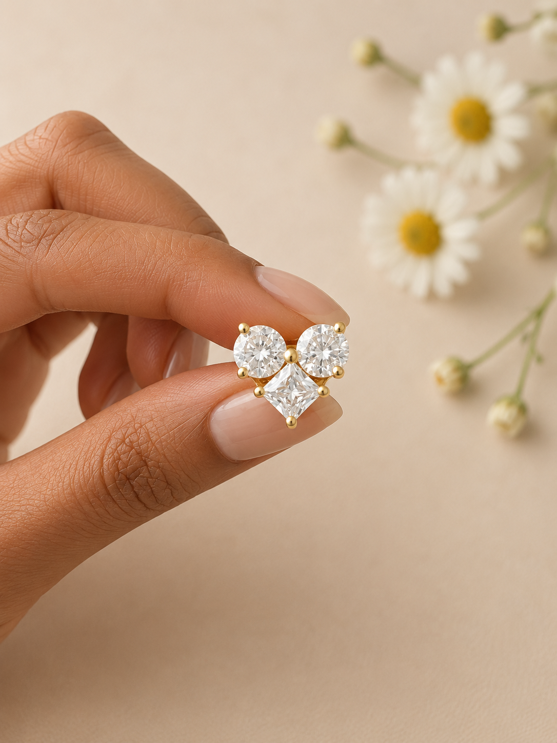 Gold earrings with clear gemstones held by a hand against a beige background with flowers.
