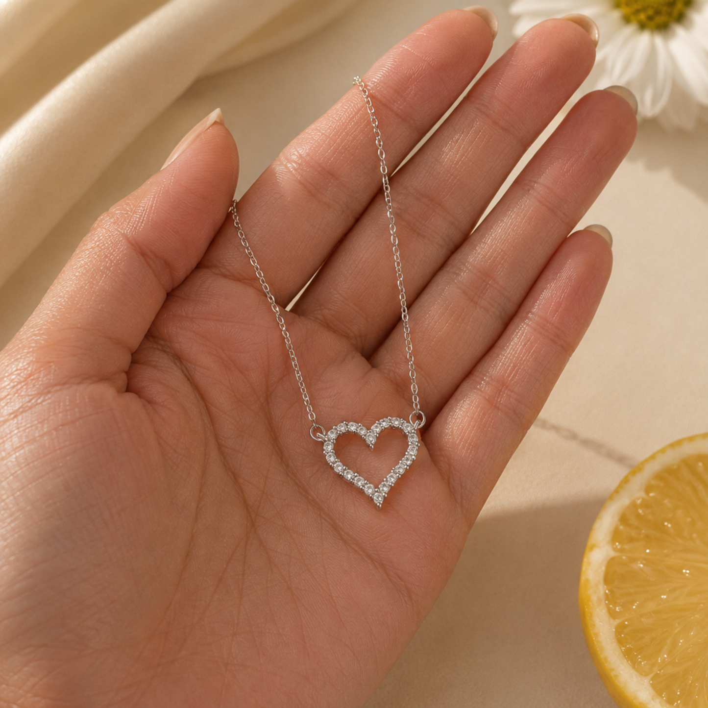 Hand holding a silver necklace with a heart pendant, with a lemon and white flower in the background.