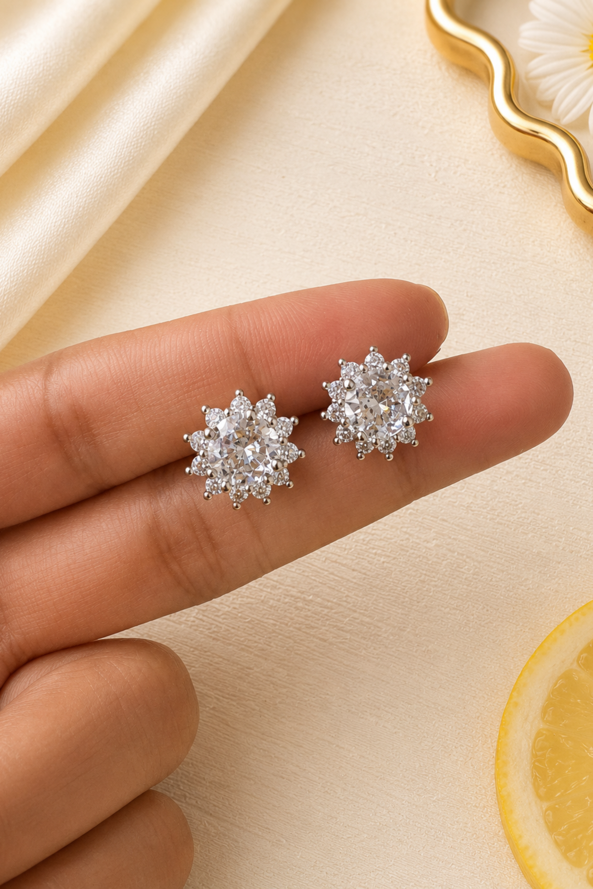 A close-up of a hand holding a pair of silver-toned stud earrings with clear gemstones in a flower-like cluster setting, against a soft cream background with decorative fabric, a daisy, and a lemon slice.