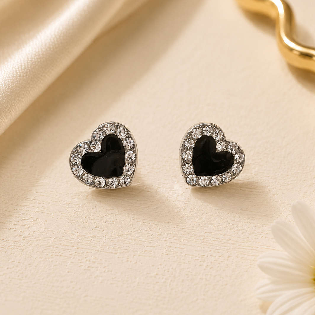 Heart-shaped earrings with black centers and clear stones on a beige fabric background with a gold ring and white flower.