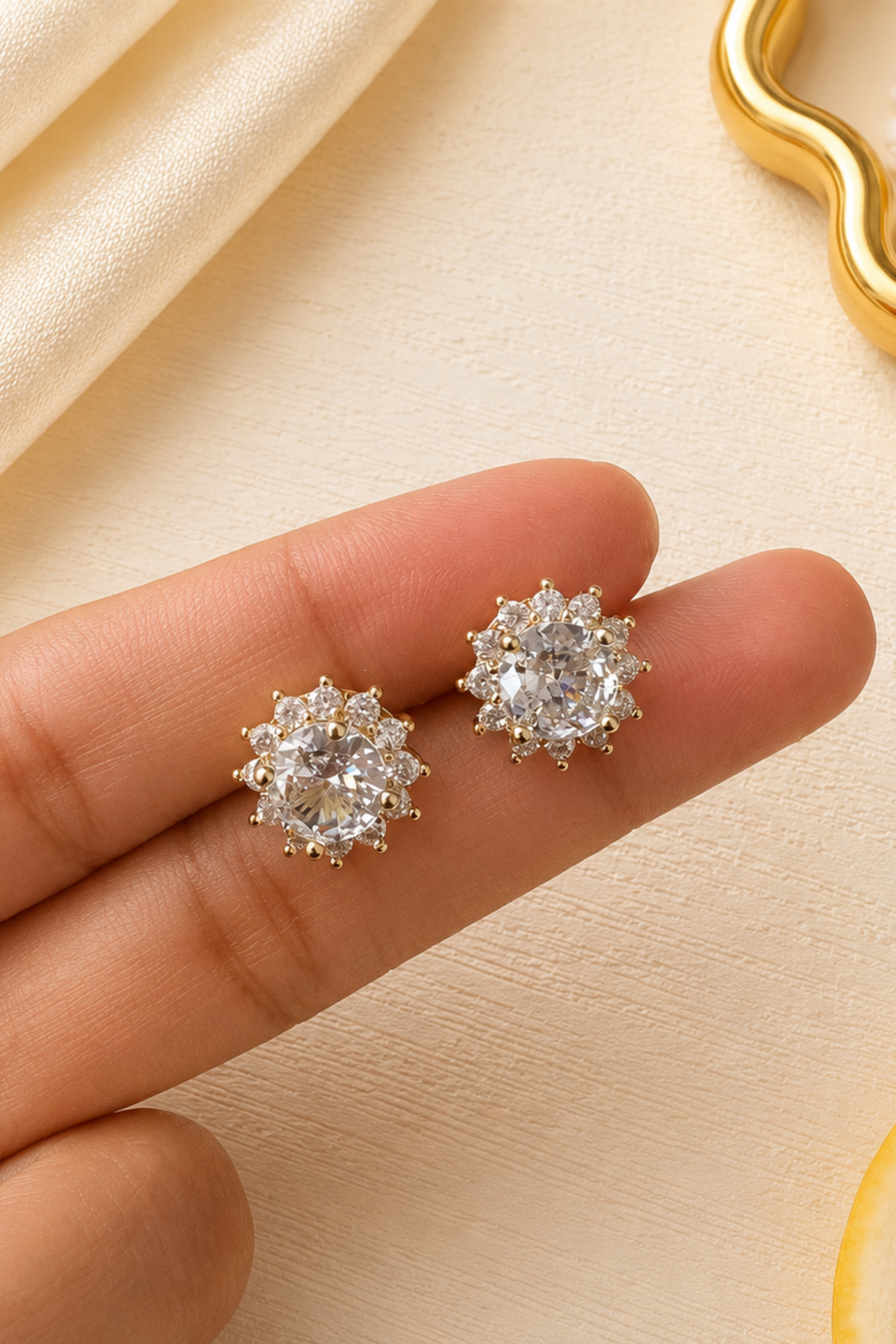 A pair of gold-toned stud earrings with clear gemstones arranged on a person's fingers, against a soft cream fabric background with decorative props.